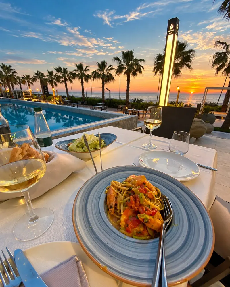 Seafood linguine and a glass of white wine on a table by the pool in Golem, palm trees and the sea at sunset behind