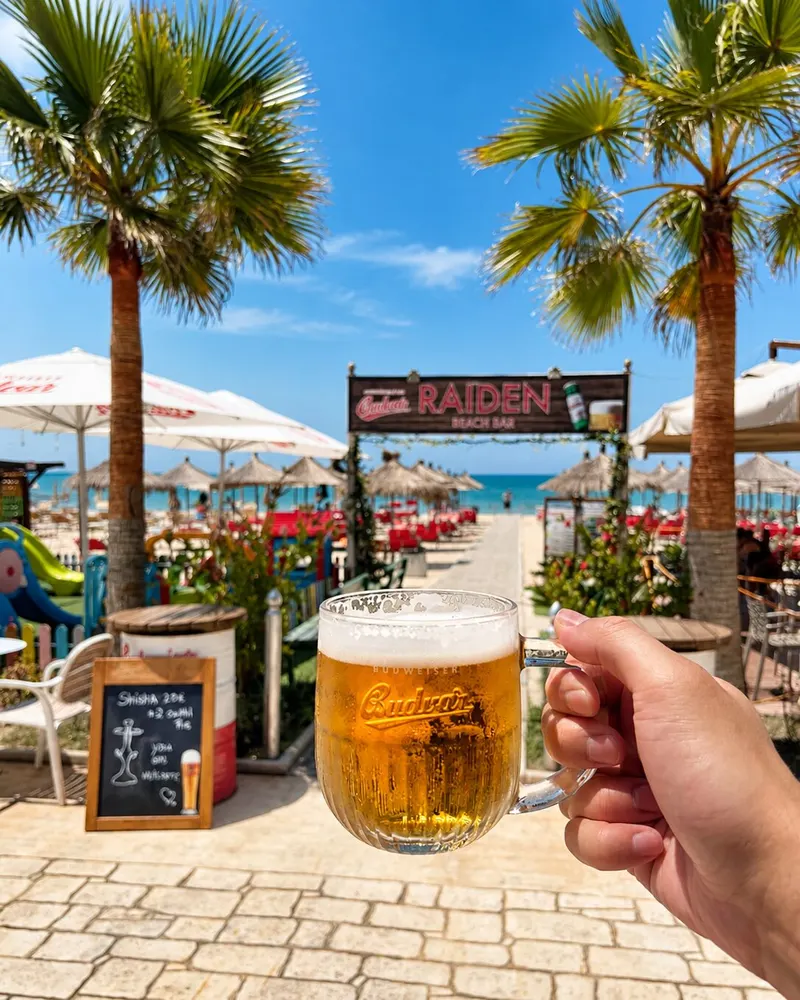 A glass of Budvar held up in front of the Beach Bar Raiden sign, palm trees and the beach behind