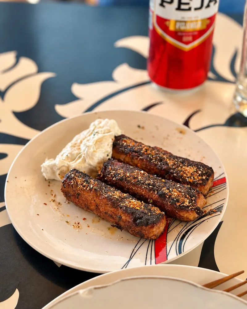 A plate of grilled qofte with a yoghurt dip next to a can of Peja beer on a restaurant table