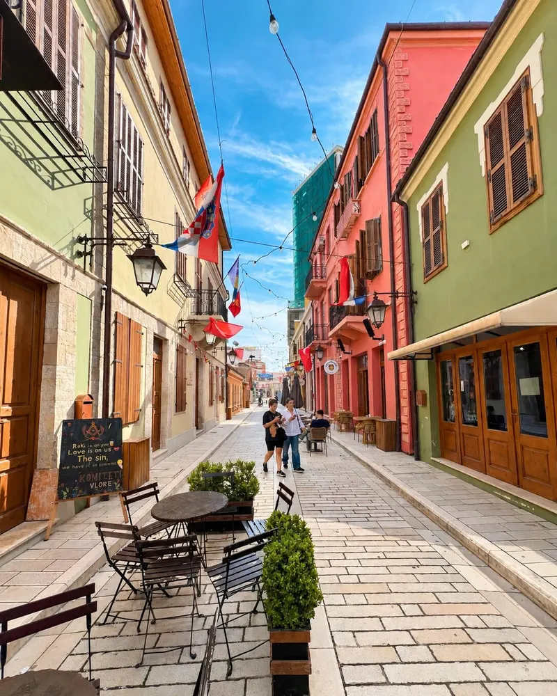 A narrow pedestrian street in Vlorë lined with pastel buildings, café tables and flags strung overhead
